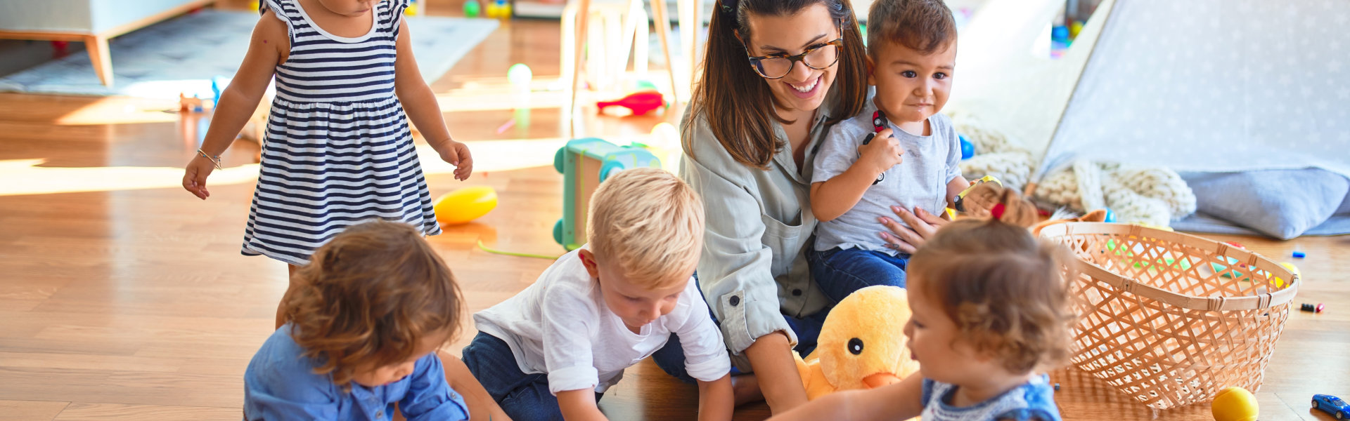 Beautiful teacher and group of toddlers sitting on the floor drawing using paper and pencil around lots of toys at kindergarten